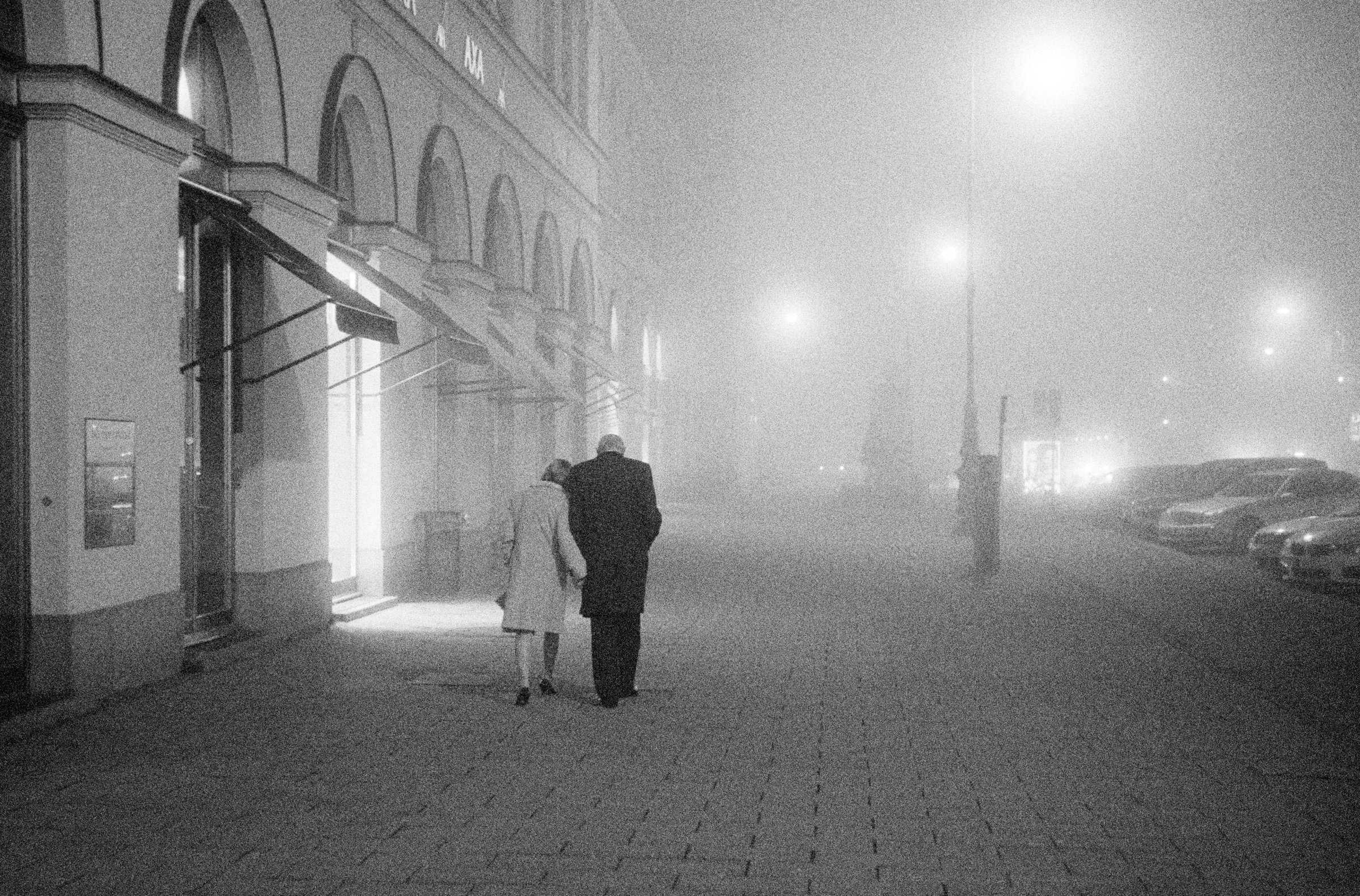 Analoge Schwarzweißfotografie von Claudius Schutte: Ein älteres Paar geht Arm in Arm durch den nächtlichen Nebel am Münchner Odeonsplatz, Straßenlaternen leuchten diffus.