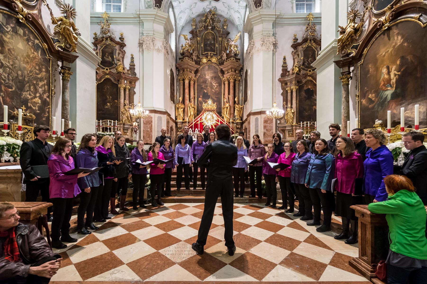 Der KammerChor KlangsCala singt im Halbkreis vor einem barocken Hochaltar, der Dirigent steht mit dem Rücken zur Kamera